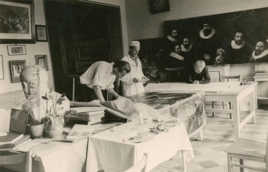 Melnbalts fotoattēls, kurā redzamas vairākas personas, kas iesaistītas dažādās aktivitātēs telpā, kas piepildīta ar mākslas darbiem un klasisku dekoru. Viens cilvēks uz galda stiepj lielu auduma gabalu, bet cits raksta vai zīmē pie atsevišķa galda. Telpu 